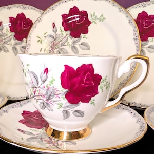 Puede incluir: Juego de taza y platillo blancos con un vibrante diseño de rosa roja. La taza y el platillo tienen ribete dorado, y el juego incluye platos a juego. El patrón floral presenta hojas y capullos grises.