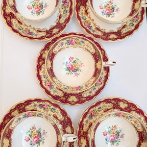 Peut inclure: Cinq tasses et soucoupes à thé vintage. Chaque tasse et soucoupe présente un intérieur blanc avec un motif de bouquet floral. Les bords sont bordeaux foncé avec des accents dorés. Les soucoupes ont un bord festonné.