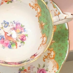Puede incluir: Juego de taza y platillo de té vintage. La taza y el platillo de porcelana blanca presentan diseños florales y de pájaros en rosa, azul y amarillo. Los bordes están adornados con oro y la taza tiene un borde verde con detalles dorados.