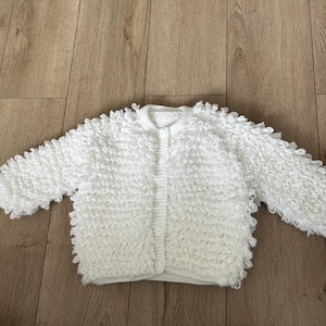 Könnte beinhalten: Eine weiße Langarmstrickjacke mit einem strukturierten Schlaufenmuster auf dem gesamten Körper und den Ärmeln. Das Kleidungsstück verfügt über einen Rundhalsausschnitt und eine vertikale Knopfleiste vorne mit vier passenden weißen Knöpfen.