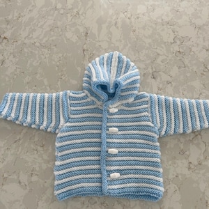 Op de afbeelding: Een gebreid babyvest met blauwe en witte strepen, een capuchon en witte knopen.