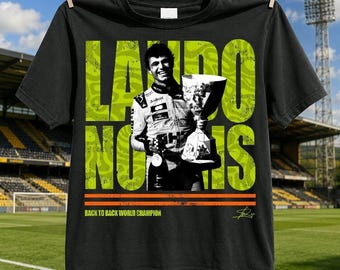May include: A black t-shirt featuring a graphic of Lando Norris holding a trophy. The text "LANDO NORRIS" appears in large lime green letters behind the figure. Below, two orange horizontal lines sit above the text "BACK TO BACK WORLD CHAMPION".