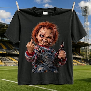 Peut inclure: Un t-shirt noir à manches courtes avec une impression graphique représentant une poupée aux cheveux orange vif, au visage cicatrisé et portant une salopette en jean bleu. La poupée tient une bouteille en verre marron et fait un doigt d'honneur.