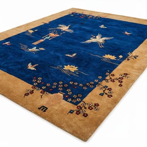 Pode incluir: Um tapete retangular com um campo central azul profundo apresentando motivos bordados de grous brancos, borboletas e flores de lótus. O tapete tem uma borda larga em tom castanho decorada com ramos floridos e um vaso no canto.