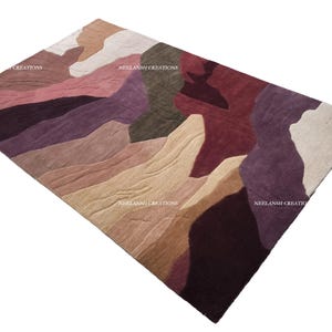May include: A rectangular area rug featuring an abstract pattern of organic, flowing shapes in shades of burgundy, deep purple, olive green, beige, and soft pink. The text NEELANSH CREATIONS is visible in multiple locations across the rug surface.