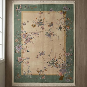 May include: A rectangular rug with a cream-colored center and a green border. The rug features a floral design with pink, purple, and green flowers and branches. The rug has a fringed edge.