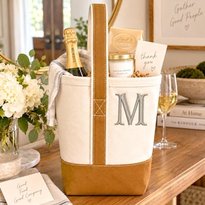 Op de afbeelding: Een crèmekleurige canvas tas met een bruine onderkant en handvat, voorzien van een geborduurde grijze letter M op de voorzijde. De tas bevat een fles Korbel-champagne, een doos Lindt Lindor-chocolaatjes, een Sandalwood Vanilla-kaars en een bedankkaartje.