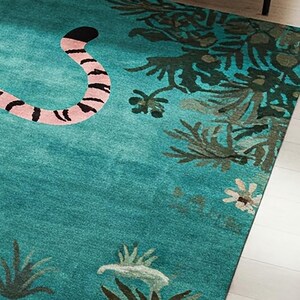 May include: A teal area rug featuring a pink and black striped animal tail on the left side, with dark green botanical foliage and small white flowers printed along the right edge and bottom border of the rectangular floor covering.