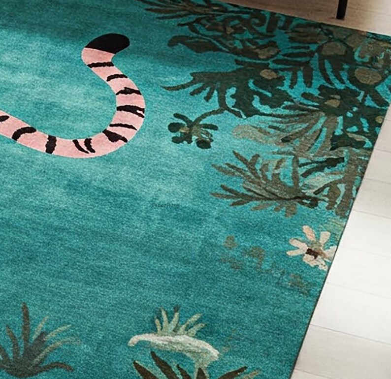 May include: A teal area rug featuring a pink and black striped animal tail on the left side, with dark green botanical foliage and small white flowers printed along the right edge and bottom border of the rectangular floor covering.