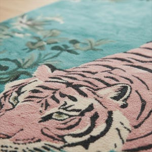 May include: A tufted rug featuring a large pink tiger with dark grey stripes positioned against a vibrant teal background. The design includes detailed floral elements in shades of green and white, showcasing a textured pile surface.