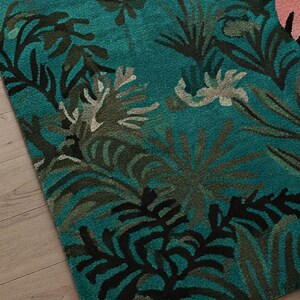 May include: A tufted area rug featuring a deep teal background with a dense pattern of dark green, black, and light gray tropical palm leaves and foliage. A portion of a light pink section with black line details is visible on the right side.