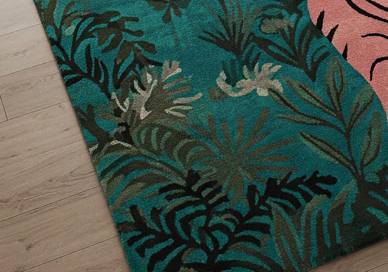 May include: A tufted area rug featuring a deep teal background with a dense pattern of dark green, black, and light gray tropical palm leaves and foliage. A portion of a light pink section with black line details is visible on the right side.