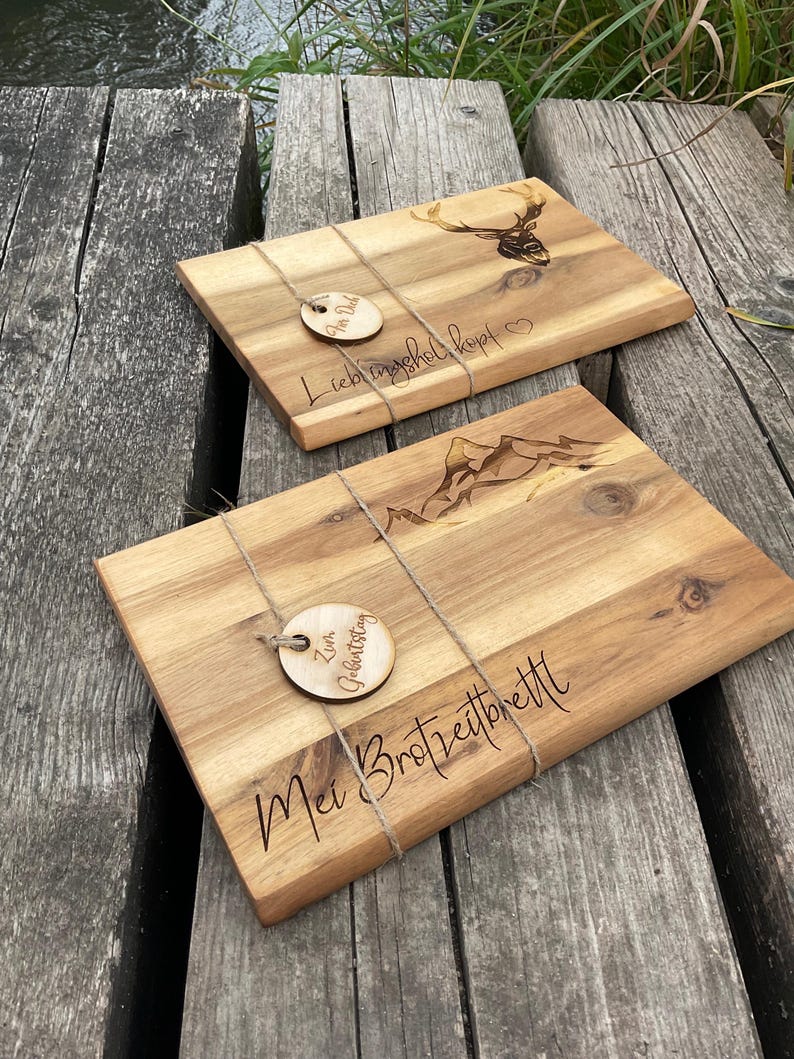 May include: Two rectangular wooden chopping boards with engraved designs. One has a deer head, the other mountains. Each board is tied with twine and has a small wooden tag with text. The boards are on a weathered wooden surface.