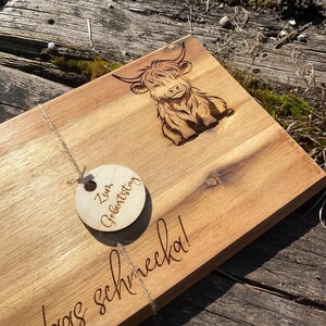 May include: Wooden serving board with a carved Highland cow design. Includes a small wooden tag with the words "Zum Geburtstag" and the phrase "was schmecka!" etched into the wood. The board is made of light brown wood.