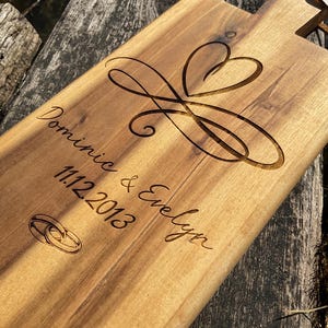 Może przedstawiać: Drewniana deska do krojenia z wygrawerowanym sercem i dwoma pierścieniami. Na desce wygrawerowany jest również tekst "Dominie & Evelyn 11.12.2013".