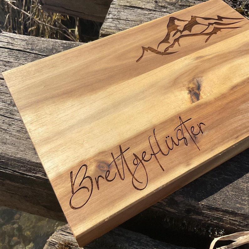 May include: A rectangular wooden chopping board with a mountain range design etched into the top. The words "Brettgefl&uuml;ster" are also etched into the wood. The board is light brown and appears to be made of acacia wood.