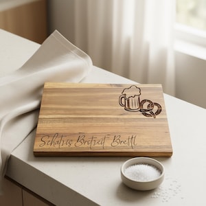 May include: Wooden chopping board with a beer mug and pretzel design. The board has the text "Schatz Brotzeit Brett" engraved on it. A small bowl of salt sits in front of the board.