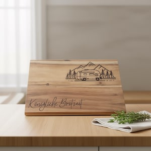 May include: Wooden chopping board featuring a campervan and mountain design. The board is engraved with the words "Konigliche Brotzeit". A sprig of rosemary and a white napkin are on the table.