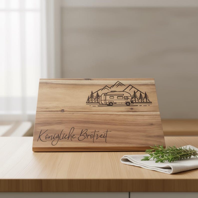 May include: Wooden chopping board featuring a campervan and mountain design. The board is engraved with the words "Konigliche Brotzeit". A sprig of rosemary and a white napkin are on the table.