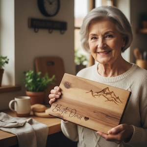 May include: A rectangular wooden chopping board featuring a mountain range design and the words "Beste Oma" and a heart. The board is made of light-coloured wood.