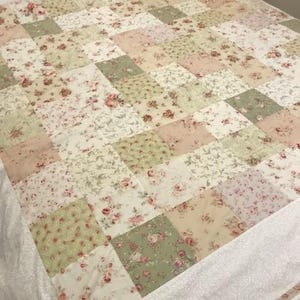 Op de afbeelding: Een patchwork quilt met een bloemenpatroon in tinten roze, groen en crème. De quilt is gemaakt van vierkante stoffen lappen en heeft een witte rand. De quilt ligt op een bed.