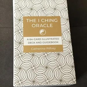 May include: A deck of oracle cards titled "The I Ching Oracle" by Catherine Pilfrey. The box has a gold and white geometric pattern. The title is in gold lettering on a gold background, with a yin-yang symbol.