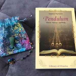 May include: A green pendulum with a chain and a small bag. The book in the background is titled "Pendulum: Oracle - Advice - Self-help" by Susanne Peymann. The book is part of the "Library of Oracles" series.