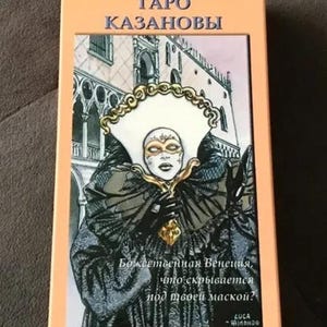 May include: A tarot card deck box featuring a colourful illustration of a masked figure in front of a building. The text on the box reads "Taro Kazanovy" and "Bozhestvennaya Venetsiya, chto skryvaetsya pod tvoe maskoy?"