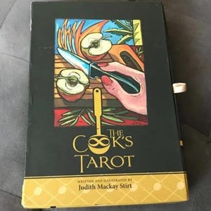 May include: A black box with the title "The Cook's Tarot" in gold lettering. The cover art features a hand holding a knife cutting an apple, with other apple slices and cooking-related imagery. The author's name is also printed on the box.