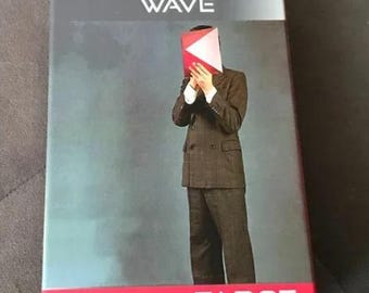May include: A tarot card box with the words "THE NEW WAVE" in white text on a gray background. The box has a red border and the word "TAROT" in white. A person in a suit holds a red and white object in front of their face.