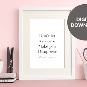 May include: A white framed print with a black and white quote that reads "Don't let anyone make you disappear - Heartstopper". The print is on a pink surface with a potted succulent plant, a black clothespin, and a white pencil holder with pencils and scissors.