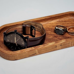 Può includere: Un vassoio di legno con un orologio e due anelli. Il vassoio è di forma ovale ed è fatto di legno marrone chiaro. L'orologio ha un quadrante nero e un cinturino in pelle marrone. Gli anelli sono d'oro e d'argento.