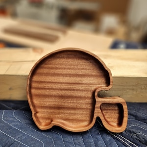May include: A wooden serving tray shaped like a football helmet. The tray is made of dark brown wood with a smooth finish.