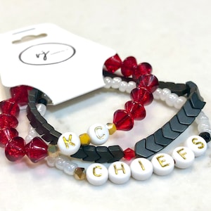 Könnte beinhalten: Drei Perlenarmbänder mit dem Wort "CHIEFS" in weißen Perlen mit goldenen Buchstaben. Die Armbänder sind mit roten, weißen und schwarzen Perlen geschichtet.