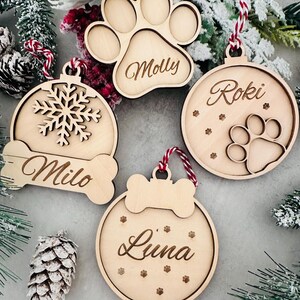 Könnte beinhalten: Vier Weihnachtsornamente aus Holz zum Thema Haustiere. Eines ist wie ein Pfotenabdruck mit dem Namen "Molly" geformt. Ein weiteres ist eine Schneeflocke mit "Milo". Das dritte ist ein Kreis mit "Roki" und Pfotenabdrücken. Das letzte ist ein Kreis mit "Luna" und Pfotenabdrücken.
