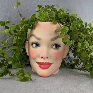 Könnte beinhalten: Ein Keramik-Pflanztopf in Form eines Frauenkopfes mit blauen Augen, roten Lippen und einem zarten rosa Rouge. Der Topf ist mit grünen, blättrigen Pflanzen gefüllt.