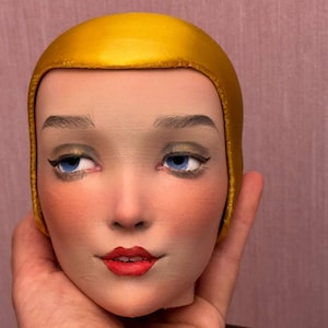 Può includere: Una testa di bambola in porcellana con un berretto in raso dorato. La bambola ha gli occhi azzurri, le labbra rosse e un viso dipinto.