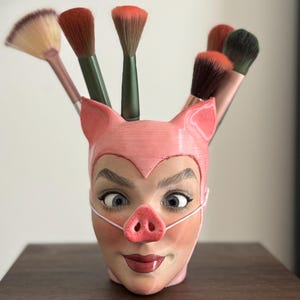 Könnte beinhalten: Ein rosafarbener, keramik-Makeup-Pinselhalter in Form eines Schweins mit einem weißen Gesicht und einer rosa Schnauze. Fünf Makeup-Pinsel ragen aus der Oberseite heraus.