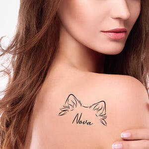 May include: A black temporary tattoo on a shoulder. The tattoo features a stylized outline of animal ears above the name "Nova" in cursive script. The skin is fair-toned, and the background is blurred.