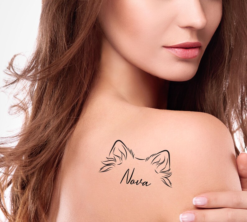May include: A black temporary tattoo on a shoulder. The tattoo features a stylized outline of animal ears above the name "Nova" in cursive script. The skin is fair-toned, and the background is blurred.