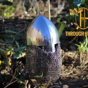 May include: A silver helmet with a chainmail face mask. The helmet is sitting on the ground in a grassy area.
