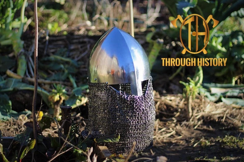 May include: A silver helmet with a chainmail face mask. The helmet is sitting on the ground in a grassy area.