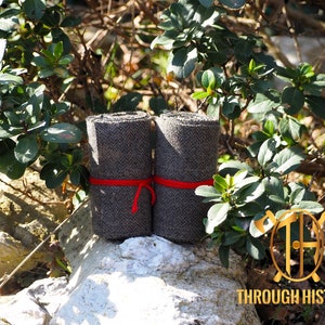 May include: Two rolls of grey herringbone fabric, bound with red twine, are presented outdoors. The fabric is tightly rolled, highlighting its texture and pattern. The scene is set amongst green foliage and natural light.
