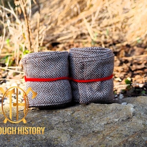 May include: Two rolled fabric items with a brown and white herringbone pattern, secured with a red band. The items are resting on a stone surface, with a blurred natural background. The logo "THROUGH HISTORY" is visible.