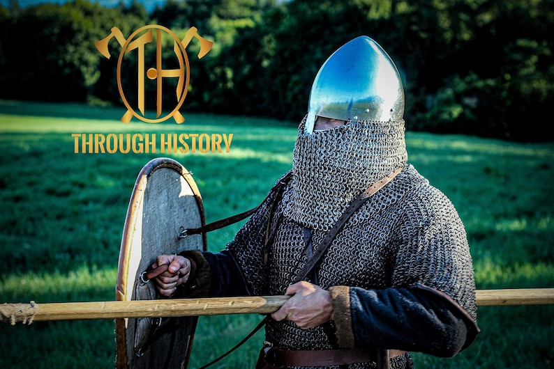 May include: A man wearing a silver helmet and chainmail armor holds a wooden shield and a wooden staff. The background is a green grassy field with trees in the distance. The text "THROUGH HISTORY" is visible in the image.