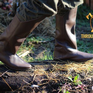 May include: A pair of brown leather knee-high boots with a pointed toe. The boots are worn by a person walking on a log in a grassy area. The text "THROUGH HISTORY" is visible in the background.