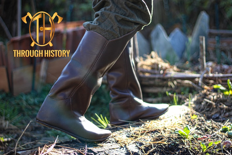May include: Brown leather knee-high boots with a pointed toe and a low heel. The boots are worn by a person standing on a wooden surface.