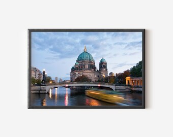 Puede incluir: Una impresión artística enmarcada que muestra la Catedral de Berlín con su cúpula verde, situada tras un puente de piedra sobre un río. Un barco amarillo borroso se mueve por el agua al atardecer, con reflejos de luces urbanas en la superficie.