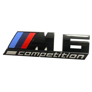 May include: Black and silver car emblem with the letters "M6 COMPETITION". The emblem has a rectangular shape with a section featuring blue, dark blue, and red stripes. The word "COMPETITION" is in silver.
