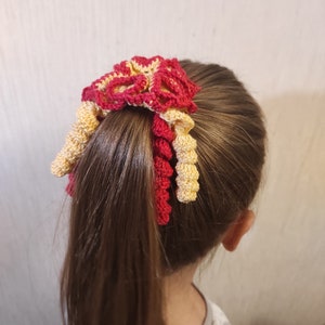 Könnte beinhalten: Ein rotes und gelbes gehäkeltes Haargummi mit Blumenmuster. Das Haargummi ist um einen Pferdeschwanz gebunden.
